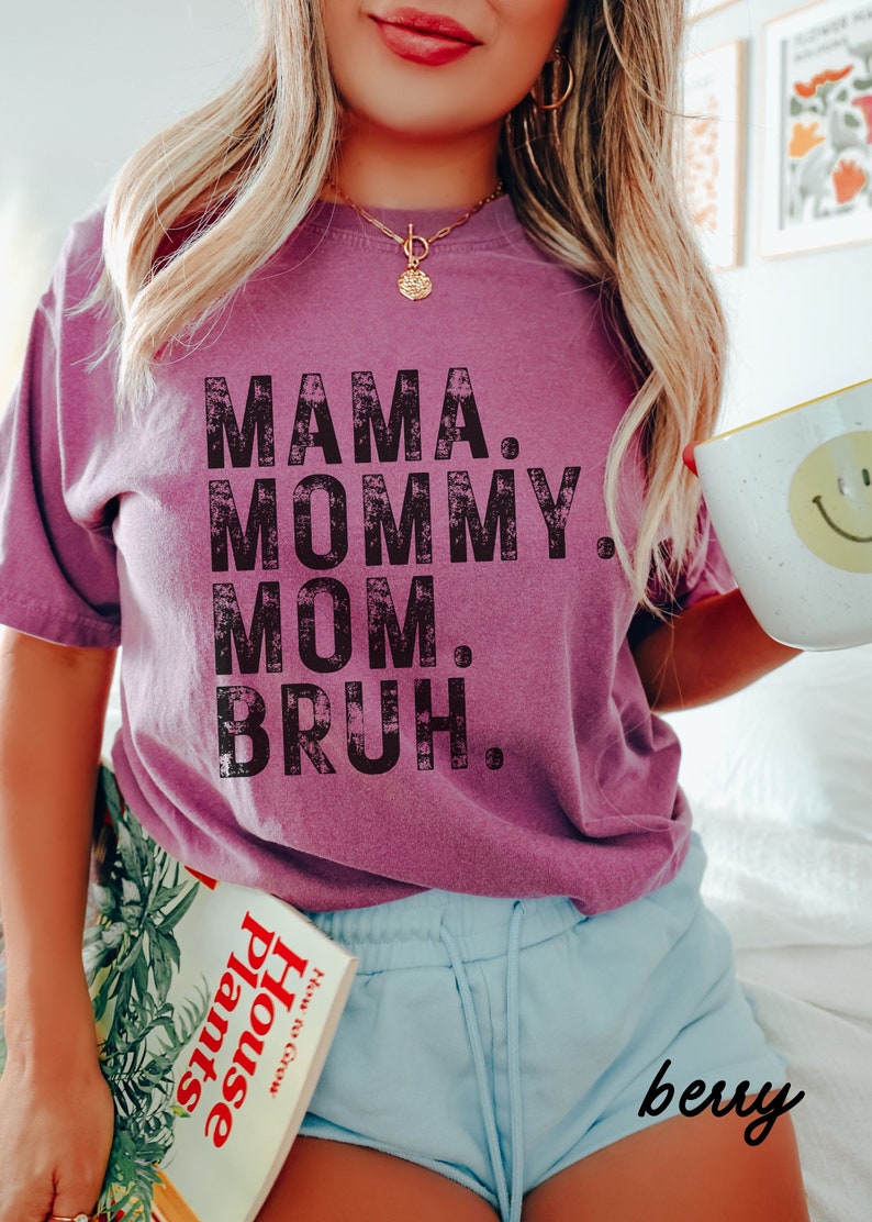 May include: A woman wearing a purple t-shirt with the text "MAMA. MOMMY. MOM. BRUH." in black distressed lettering. She is holding a mug and a book titled "House Plants".