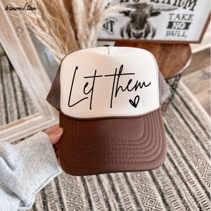 May include: A brown and tan trucker hat with the words "Let them" in black script across the front. A small heart is next to the word "them". The hat has a white front panel and a brown brim and mesh back.