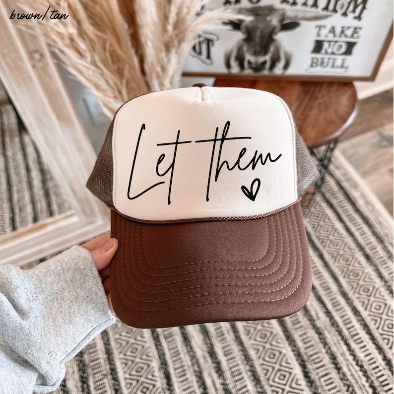 May include: A brown and tan trucker hat with the words "Let them" in black script across the front. A small heart is next to the word "them". The hat has a white front panel and a brown brim and mesh back.