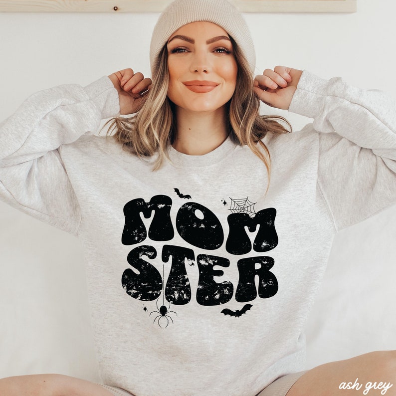 May include: A light gray sweatshirt with a black graphic that says "MOMSTER" in a retro font. The graphic also includes a spider, a spider web, and bats.