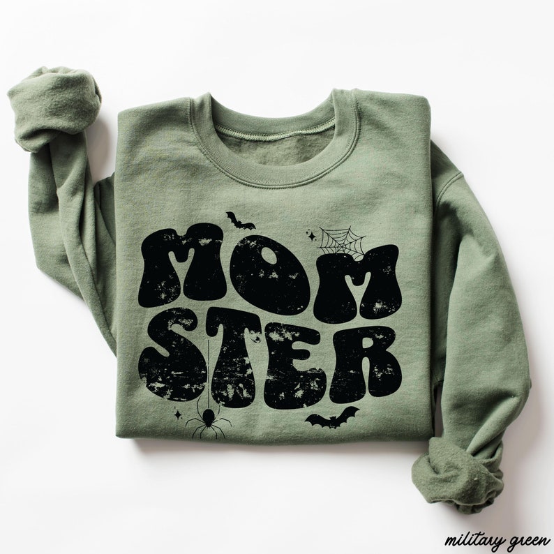 May include: A green sweatshirt with the text "MOMSTER" in black, retro-style lettering. The text is surrounded by small black spiderwebs, bats, and stars.