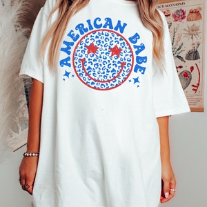 May include: White oversized t-shirt with a blue and red graphic of a smiley face with stars for eyes and the text "American Babe".