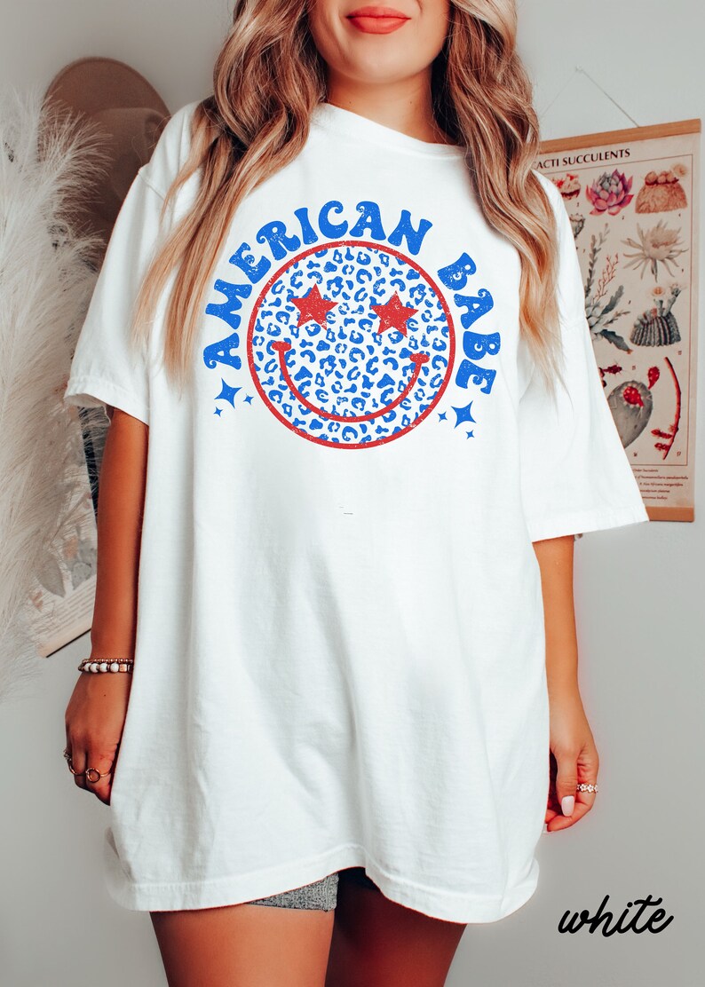 May include: White oversized t-shirt with a blue and red graphic of a smiley face with stars for eyes and the text "American Babe".