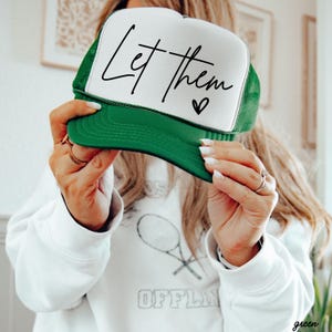 May include: A green and white trucker hat with the text "Let them" in black script and a small heart. The hat has a green brim and mesh back, and a white front panel. The hat is a casual accessory.