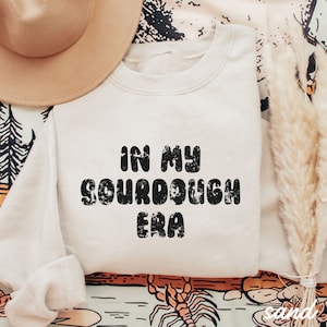 May include: A cream-colored sweatshirt with the text "In My Sourdough Era" printed in black. The text is in a retro, bubbly font.