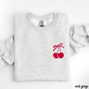 May include: Ash grey sweatshirt with a red cherry design on the front. The cherries have a red bow on top.