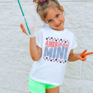May include: A young girl wearing a white t-shirt with the text "AMERICAN MINI" in pink and blue letters. She is wearing neon green shorts and has her hair pulled up in a ponytail with a pink scrunchie.
