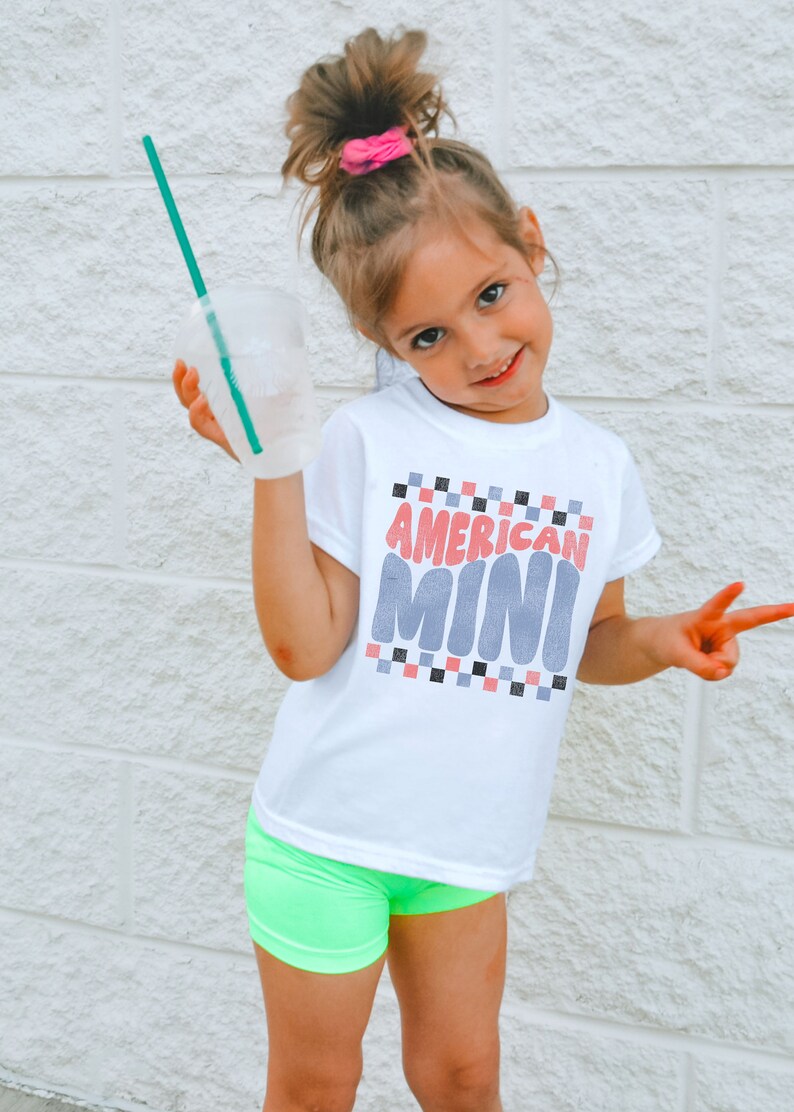May include: A young girl wearing a white t-shirt with the text "AMERICAN MINI" in pink and blue letters. She is wearing neon green shorts and has her hair pulled up in a ponytail with a pink scrunchie.