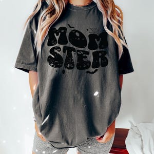 May include: A dark gray t-shirt with a black graphic that says "MOMSTER" in a retro font. The graphic also includes a spider web, bats, and stars.