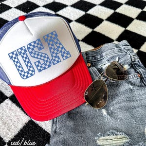 May include: A red and blue trucker hat with a white mesh back. The hat has a checkered "USA" design on the front.