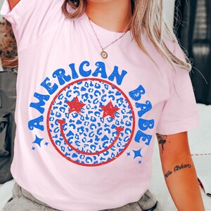 May include: Pink t-shirt with a red and blue smiley face graphic. The smiley face is filled with a blue and white leopard print pattern. The text "American Babe" is written around the smiley face.