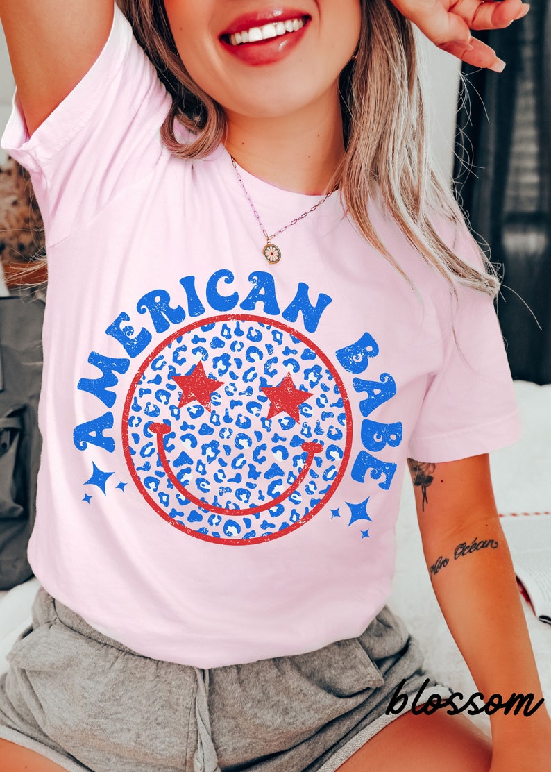 May include: Pink t-shirt with a red and blue smiley face graphic. The smiley face is filled with a blue and white leopard print pattern. The text "American Babe" is written around the smiley face.
