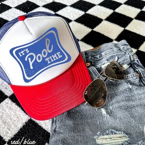 Może przedstawiać: Czapka typu trucker w kolorze czerwonym, białym i niebieskim z napisem "It's Pool Time" w niebieskim prostokącie. Czapka jest połączona z dżinsowymi szortami i okularami przeciwsłonecznymi w złotej oprawie. Tło to czarno-biały wz&oacute;r w szachownicę.