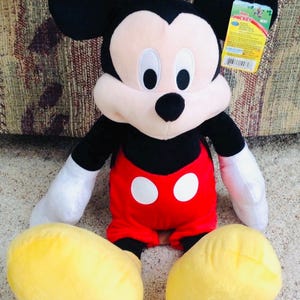 May include: A plush Mickey Mouse doll with black ears, a black nose, and a cream-colored face. Mickey wears red shorts with two white buttons, white gloves, and yellow shoes. A Disney tag is attached.