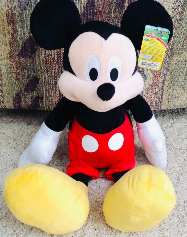 May include: A plush Mickey Mouse doll with black ears, a black nose, and a cream-colored face. Mickey wears red shorts with two white buttons, white gloves, and yellow shoes. A Disney tag is attached.