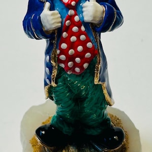 May include: A ceramic clown figurine with a red wig, a painted face, and a red and white polka-dot tie. The clown wears a blue coat with gold trim and green pants. The figurine is mounted on a white base with gold accents.