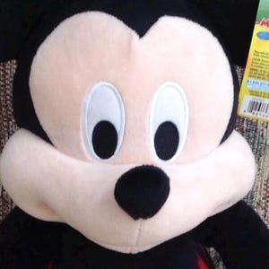 May include: A plush Mickey Mouse toy with black ears, a black nose, and a light pink face. The toy has a red body with white buttons and black arms. The background is a patterned fabric.