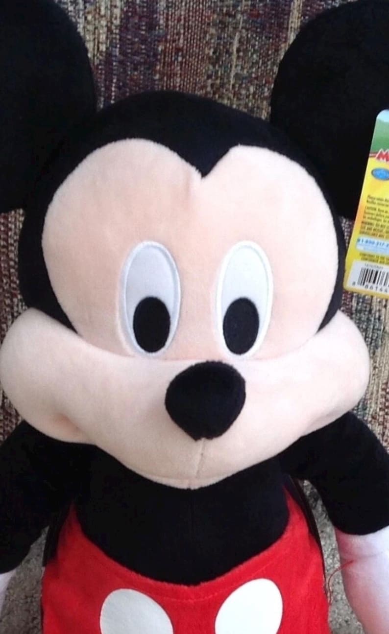 May include: A plush Mickey Mouse toy with black ears, a black nose, and a light pink face. The toy has a red body with white buttons and black arms. The background is a patterned fabric.