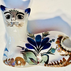 Puede incluir: Figura de gato de cerámica en posición reclinada. El gato es principalmente blanco con detalles azules en las orejas y diseños florales. El diseño floral incluye colores azul, verde y marrón. La cara del gato tiene detalles con marcas negras y marrones.