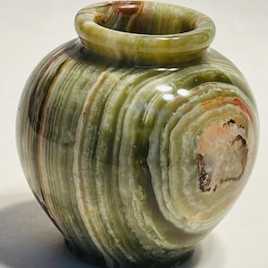 May include: A small, round vase with a wide opening, crafted from green onyx. The stone features swirling patterns of green, brown, and white, creating a natural, textured appearance. The vase has a polished, glossy finish.
