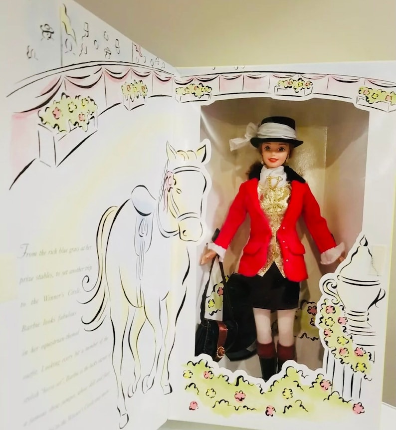 May include: A doll in a box, dressed in a red riding jacket, black skirt, and a hat with a white bow. The box has a drawing of a horse and text. The doll is holding a black purse. The background has a drawing of a horse and text.
