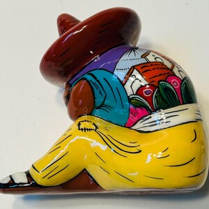 May include: A colorful ceramic figurine of a person wearing a sombrero, yellow pants, and a blue shirt. The figure is painted with bright colors and depicts a scene on the back of the figure.