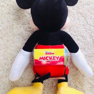 May include: A plush Mickey Mouse doll, featuring black ears, a black body, red shorts, yellow shoes, and white gloves. The doll has a Disney Junior label on its back. The toy is designed to resemble the iconic cartoon character.