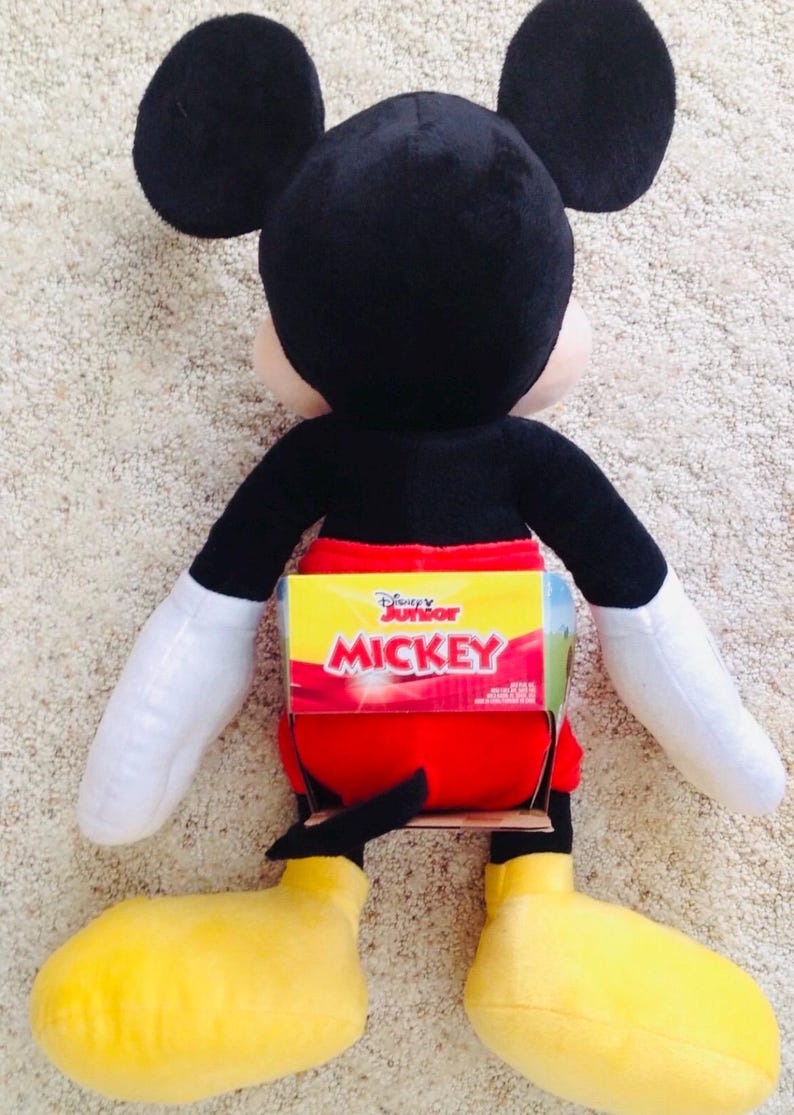 May include: A plush Mickey Mouse doll, featuring black ears, a black body, red shorts, yellow shoes, and white gloves. The doll has a Disney Junior label on its back. The toy is designed to resemble the iconic cartoon character.