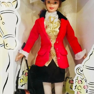 May include: A doll wearing a red jacket, black skirt, and a black hat with a white ribbon. The doll has a gold-colored vest and a black purse. The doll is in a decorative box with a horse illustration.