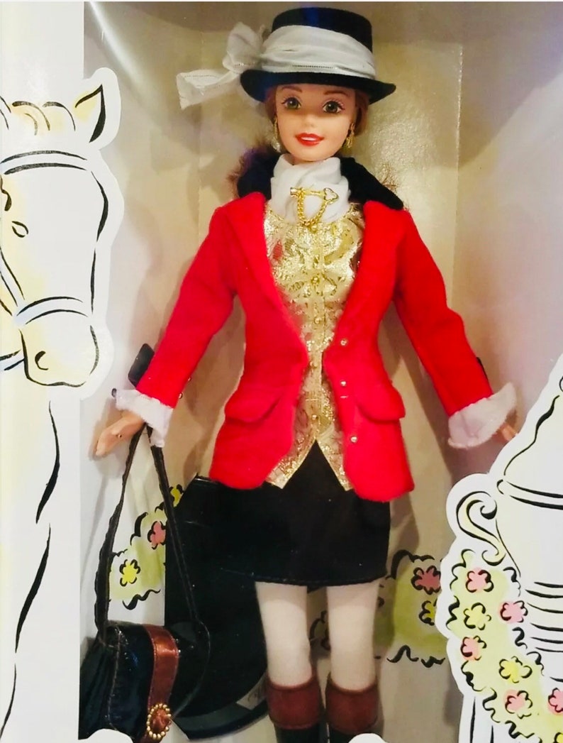 May include: A doll wearing a red jacket, black skirt, and a black hat with a white ribbon. The doll has a gold-colored vest and a black purse. The doll is in a decorative box with a horse illustration.