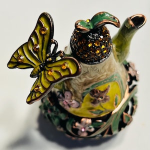 May include: A decorative teapot with a butterfly and floral design. The teapot features a yellow butterfly with brown accents and a jeweled pineapple-shaped top. The body of the teapot is green, yellow, and brown with floral and leaf accents.