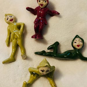 Puede incluir: Cuatro figuras de cerámica en varias poses. Una es roja con los brazos extendidos, dos son amarillas y una es verde, todas con caras sonrientes. Las figuras parecen ser vintage o antiguas.