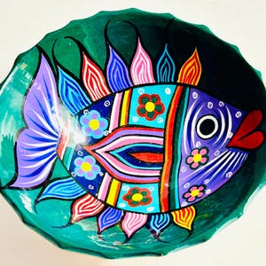 Puede incluir: Un cuenco decorativo pintado a mano con un diseño de pez colorido. El pez está pintado con vibrantes tonos de morado, azul, rojo, amarillo y rosa, sobre un fondo verde turquesa. El borde del cuenco tiene un borde festoneado.