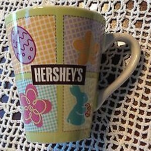 May include: A green ceramic mug with a patchwork design featuring a bunny, a flower, and an egg. The mug has a white handle and the text "HERSHEY'S" in brown letters.