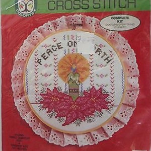 May include: A white cross stitch embroidery kit with a pink lace trim. The design features a candle with poinsettias and the words "Peace on Earth".
