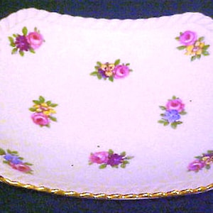 May include: A rectangular, off-white porcelain dish with a scalloped edge and gold trim. The dish is decorated with clusters of pink, purple, and yellow flowers. The flowers are arranged in a scattered pattern across the surface of the dish.