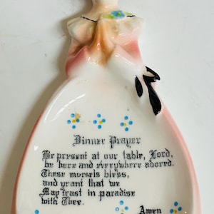 May include: A ceramic dinner prayer dish in the shape of a woman in a pink dress. The dish has a white interior with a prayer printed in black script and blue floral accents. The woman's face is painted with closed eyes.