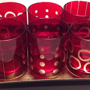 May include: Six red glass tumblers with gold rims and white geometric designs. The tumblers are arranged in a row on a gold tray.