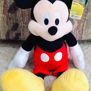 May include: A plush Mickey Mouse doll. The doll has black ears, a black head, and a white face. It wears red shorts with two white buttons, white gloves, and yellow shoes. A product tag is attached.