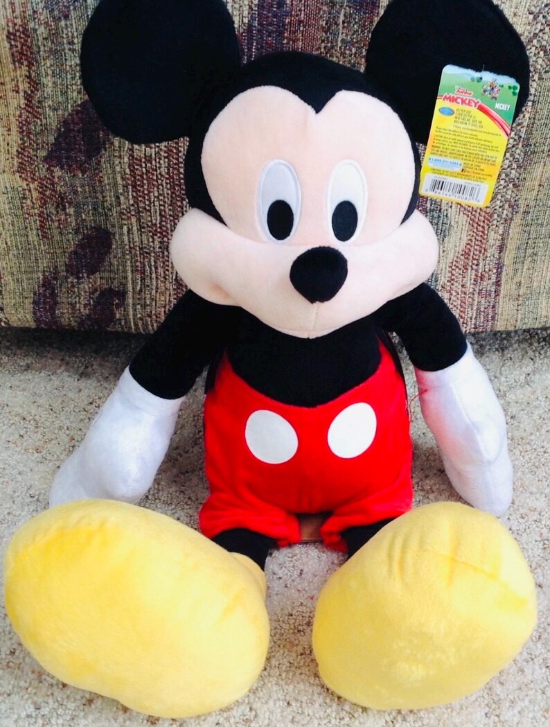 May include: A plush Mickey Mouse doll. The doll has black ears, a black head, and a white face. It wears red shorts with two white buttons, white gloves, and yellow shoes. A product tag is attached.