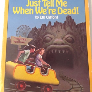 May include: A book cover with a yellow background and a cartoon illustration of two children riding a yellow roller coaster. The roller coaster is going into a dark tunnel with a scary monster's face at the entrance. The book title is "Just Tell Me When We're Dead!" by Eth Clifford.