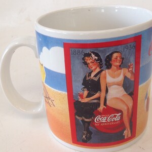 May include: White ceramic mug with a Coca-Cola 50th anniversary design. The mug features two women in vintage swimwear on a beach with a red border and the text "Drink Coca-Cola 50th Anniversary". The years 1886 and 1936 are printed on the top corners of the design.