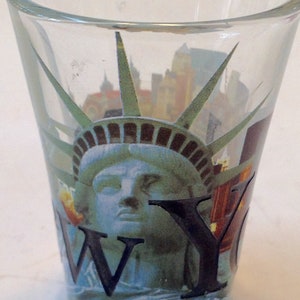 Könnte beinhalten: Ein klares Glas-Schnapsglas mit einem gedruckten Bild der Freiheitsstatue und der Skyline von New York. Das Glas hat die Buchstaben "NY" aufgedruckt.
