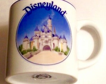 May include: White ceramic mug with a blue rim and handle. The mug features a colorful image of Sleeping Beauty's castle at Disneyland with the text "Disneyland" in blue lettering.