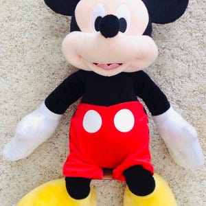 May include: A plush Mickey Mouse doll, featuring black ears, a black body, red shorts with white buttons, white gloves, and yellow shoes. The doll has a smiling face and is on a neutral-colored surface.