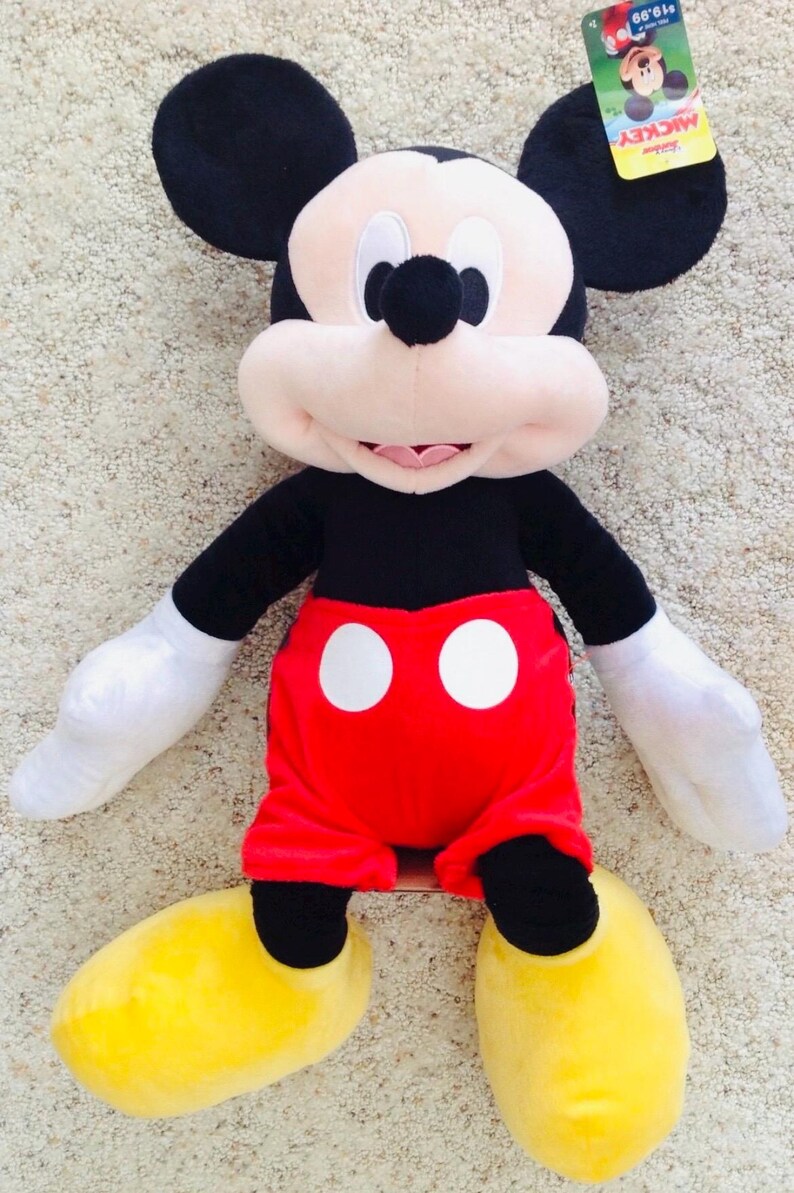 May include: A plush Mickey Mouse doll, featuring black ears, a black body, red shorts with white buttons, white gloves, and yellow shoes. The doll has a smiling face and is on a neutral-colored surface.