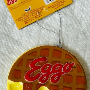 May include: A ceramic Eggo waffle ornament with a silver string for hanging. The ornament features a brown waffle design with the red "Eggo" logo and yellow syrup. The tag reads "For decoration purposes only."
