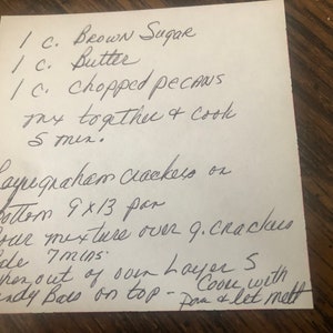 May include: A handwritten recipe on a white piece of paper, featuring instructions for a dessert. The recipe includes ingredients like brown sugar, butter, and chopped pecans. The paper is on a wooden surface.