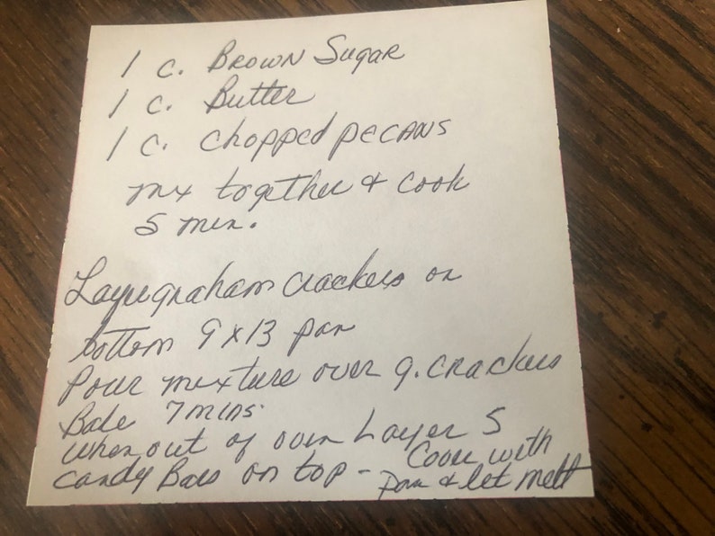 May include: A handwritten recipe on a white piece of paper, featuring instructions for a dessert. The recipe includes ingredients like brown sugar, butter, and chopped pecans. The paper is on a wooden surface.