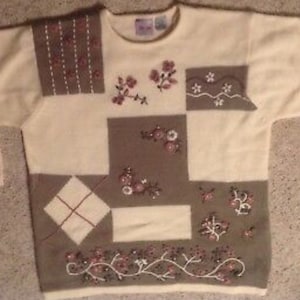 May include: A cream-colored sweater with a patchwork design featuring brown squares and embroidered floral details in shades of pink and white.